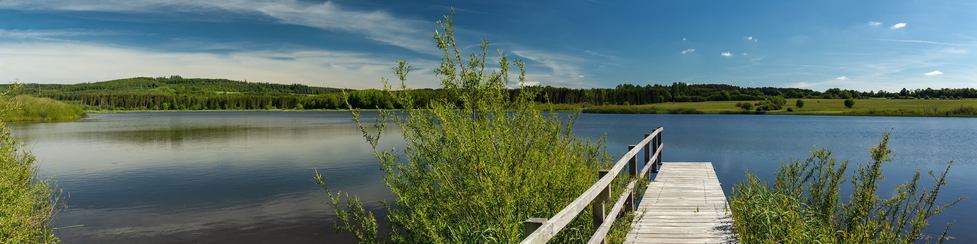 A wooden pier extending into a calm lake, surrounded by lush greenery and forested hills under a clear sky.