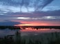 A sunset over a calm lake with reflections of the sky, surrounded by reeds and flowers.