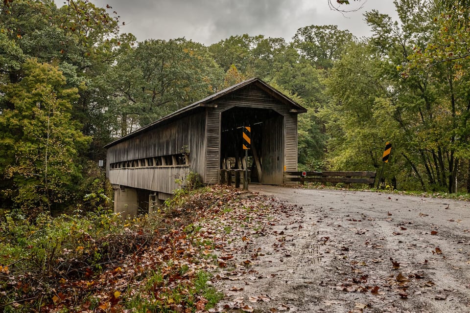 A wooden covered bridge in a forested area with a dirt path leading up to it.