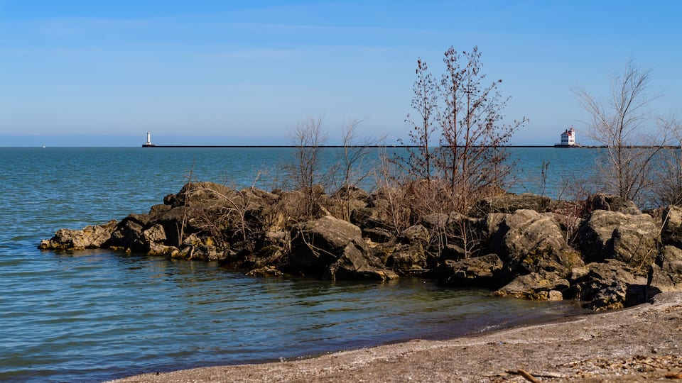 A rocky shoreline with sparse vegetation and a calm sea. In the distance, two lighthouses are visible on a breakwater.