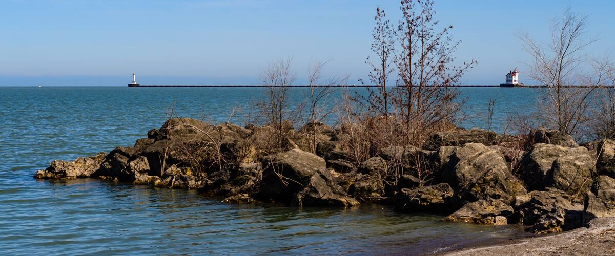 A rocky shoreline with sparse vegetation and a calm sea. In the distance, two lighthouses are visible on a breakwater.