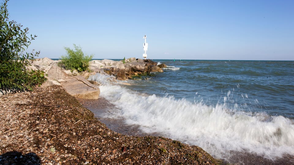 A rocky shoreline with a white lighthouse on a rock outcrop, waves crashing onto the shore, and clear blue sky.