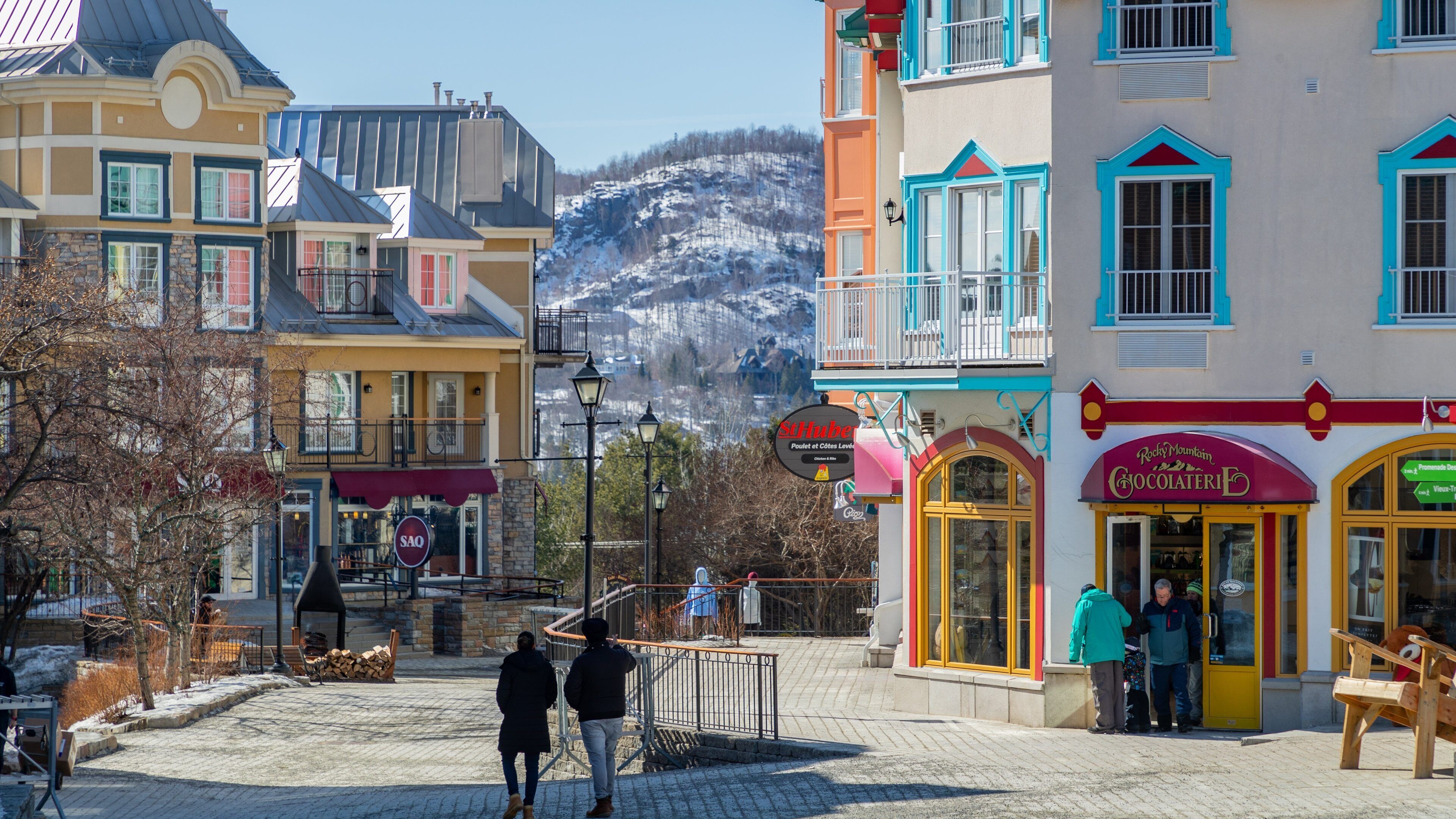 Una plaza empedrada con edificios coloridos, un letrero que indica St. Hubert, y una montaña al fondo.