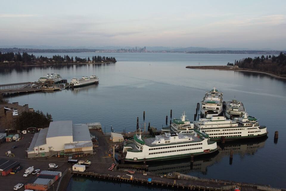 Un terminal de transbordador con un ferry grande atracado, rodeado de agua y un horizonte urbano a lo lejos.
