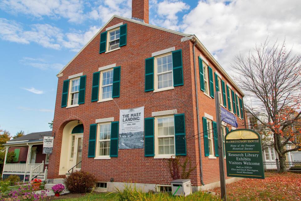 Un edificio storico in mattoni con persiane verdi, un cartello con la scritta 'The Mast Landing: Industry, Commerce, Its People 1720s-1920' e un cartello 'Barrington House' che indica la sede della Freeport Historical Society.