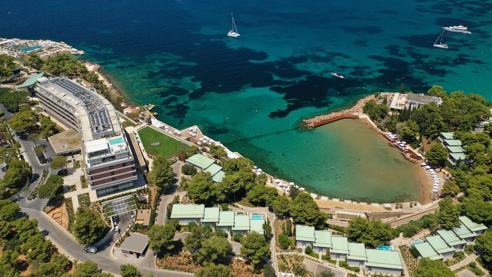 An aerial view of a coastal area with clear blue waters, a marina with boats, and a resort with green-roofed buildings.
