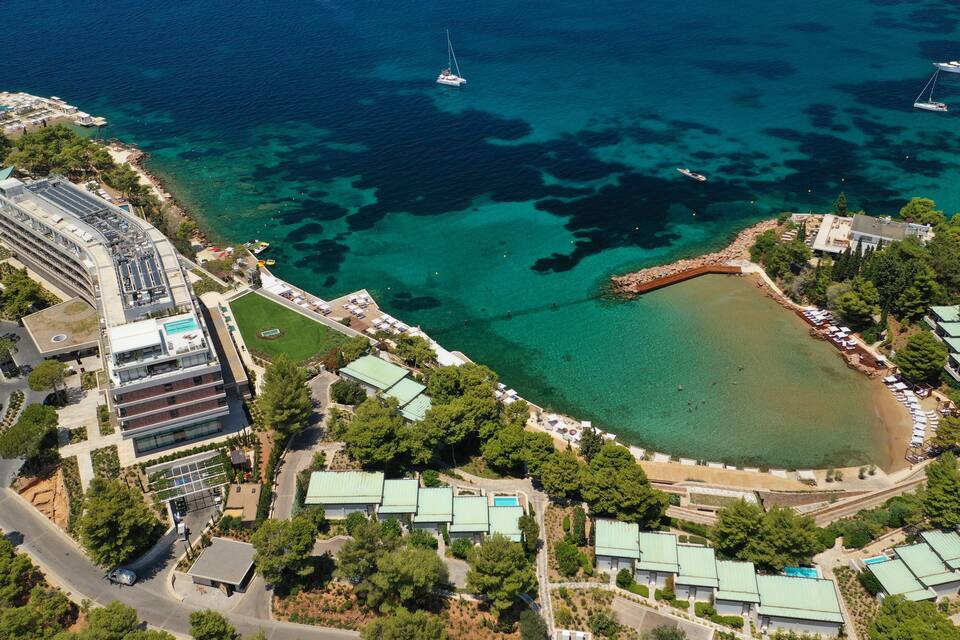 Vista aérea de uma área costeira com águas azul-claras, uma marina com barcos e um resort com edifícios de telhado verde.