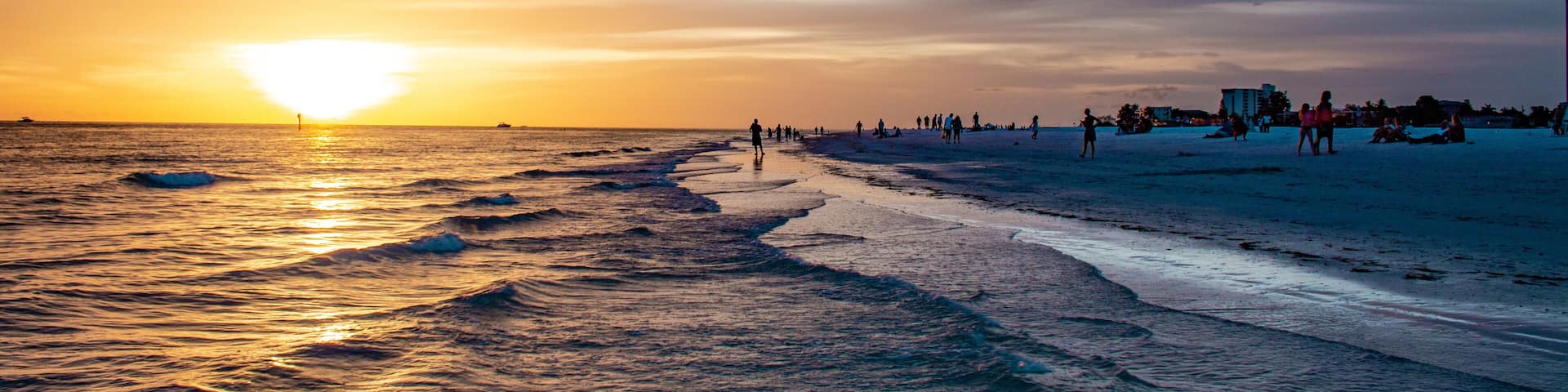 A beach at sunset with waves, people, and distant buildings.