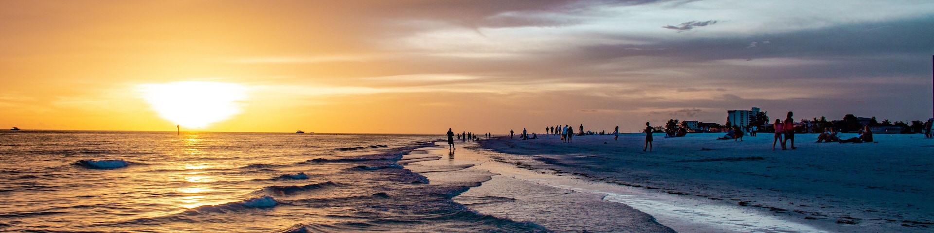 Ein Strand bei Sonnenuntergang mit Wellen, Menschen und Gebäuden in der Ferne.