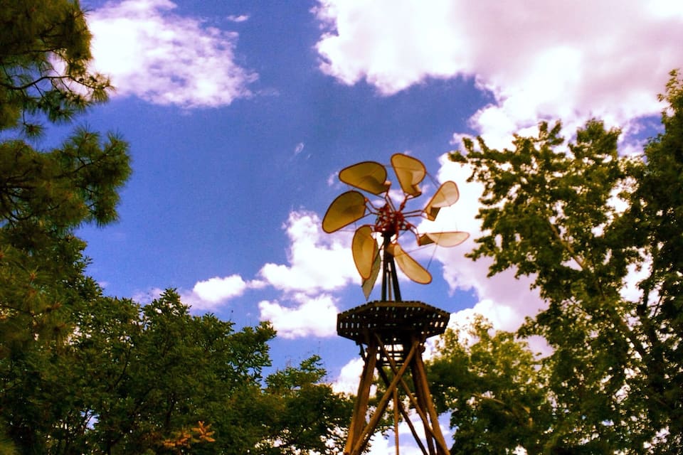 A windmill with a large fan, surrounded by trees and a blue sky with clouds.