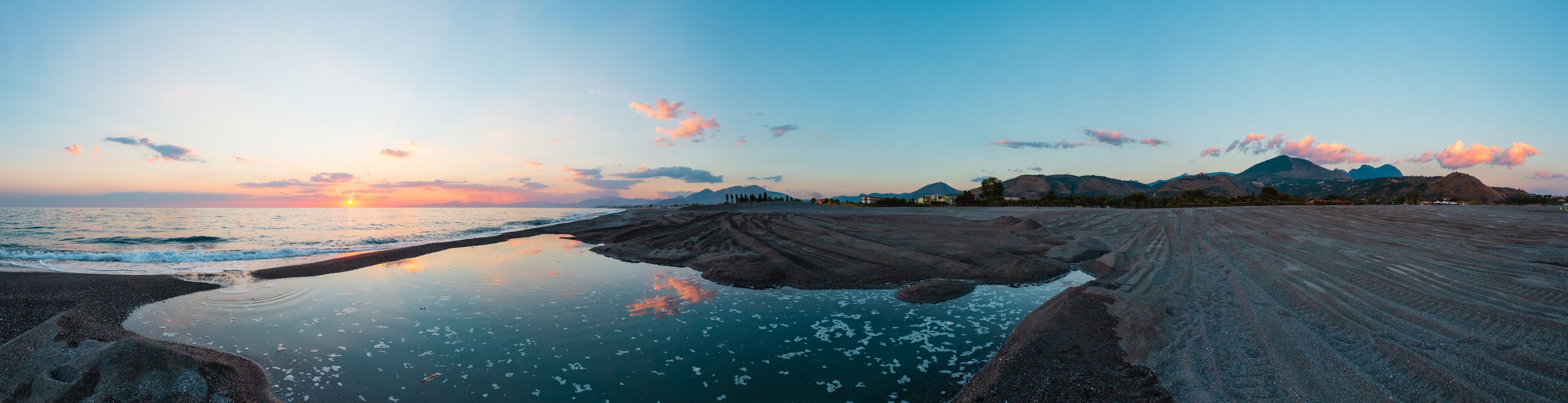A beach at sunset with a calm sea, rocky shoreline, and distant mountains.