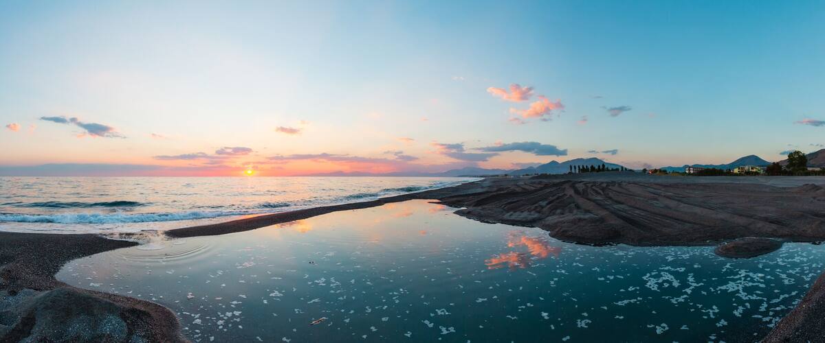 A beach at sunset with a calm sea, rocky shoreline, and distant mountains.