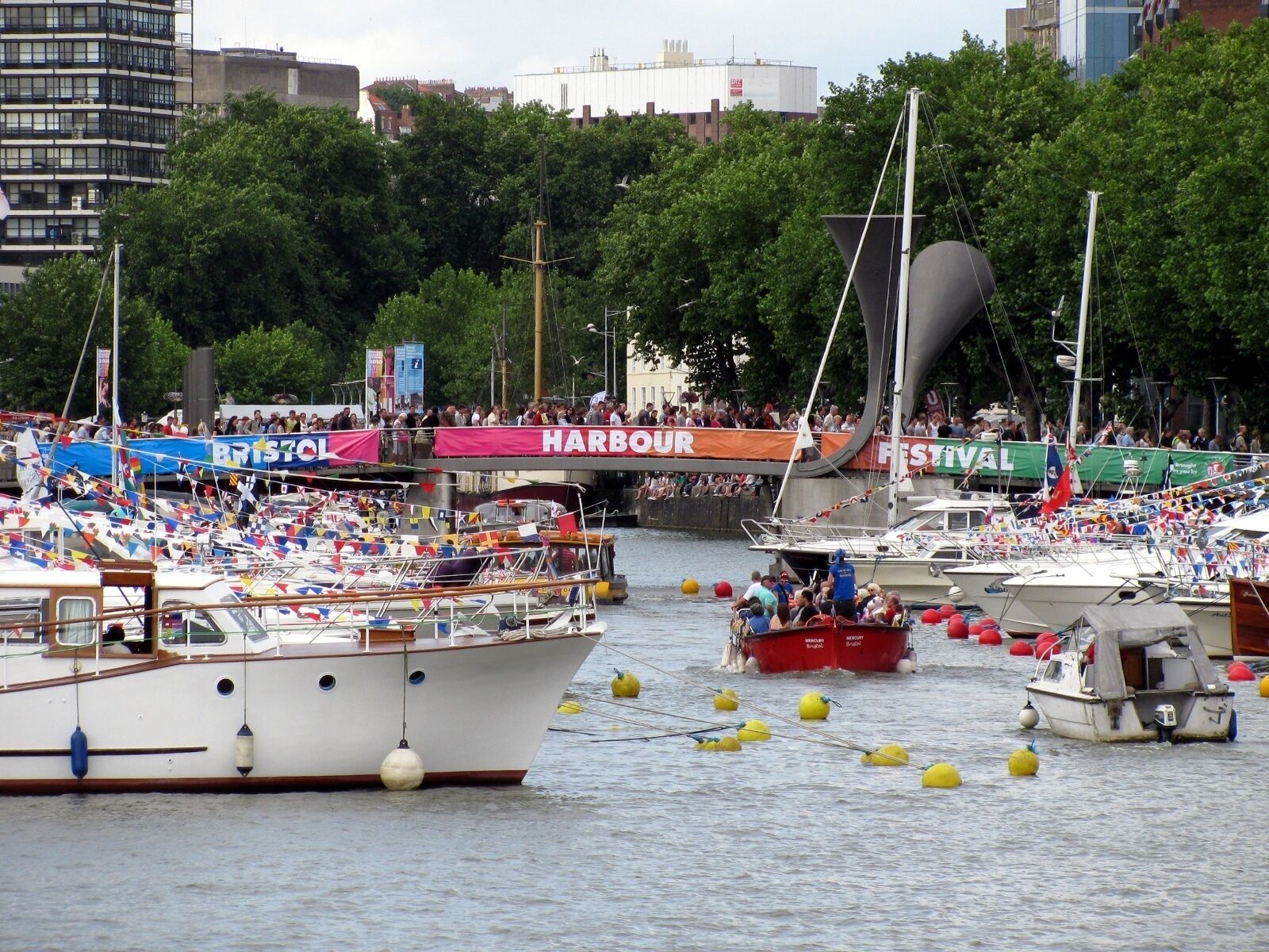 A crowded harbor with numerous boats, some with flags, and a large crowd of people watching.