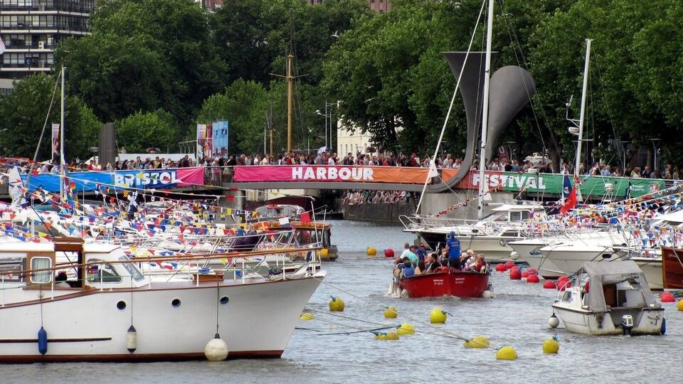 A crowded harbor with numerous boats, some with flags, and a large crowd of people watching.