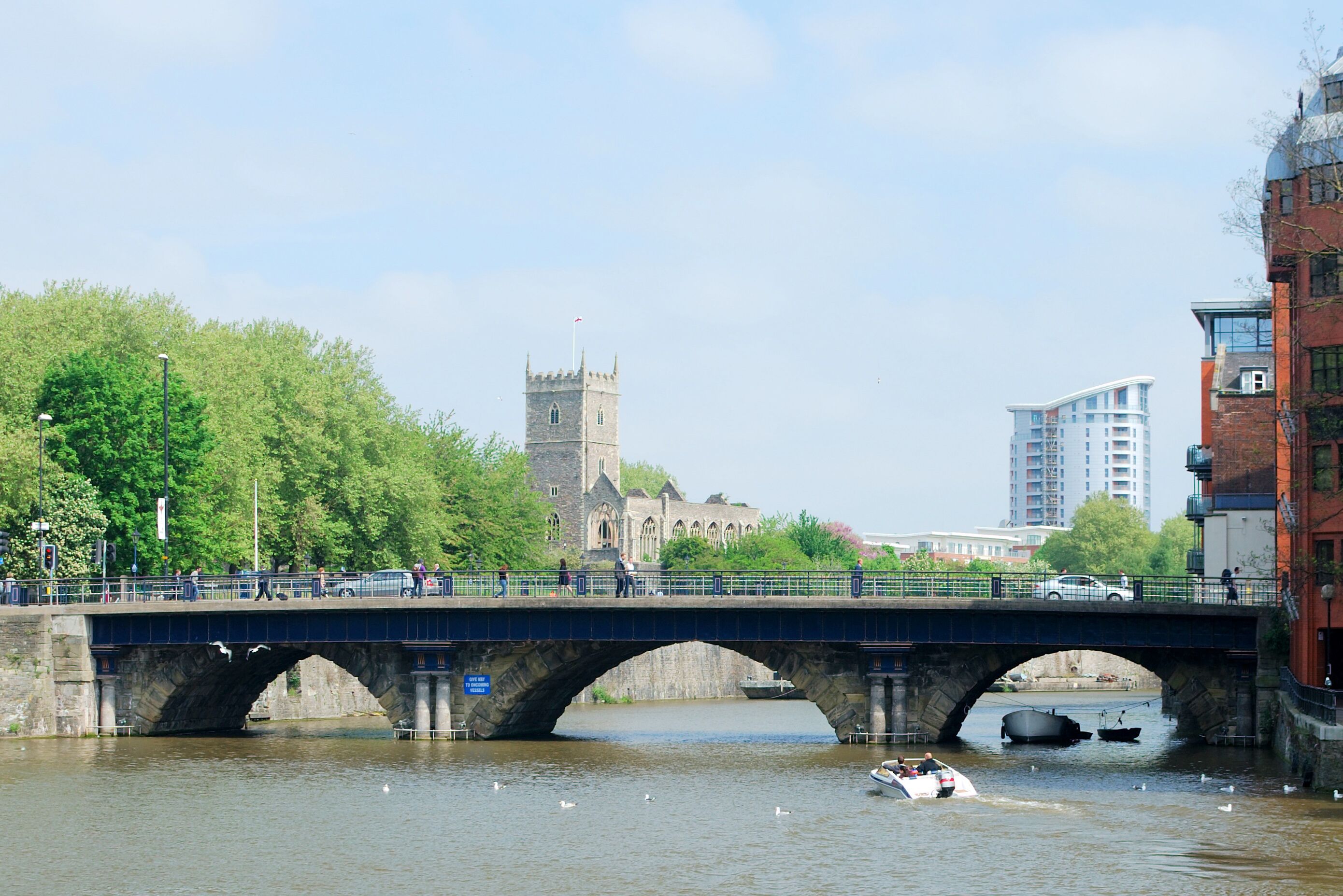 A stone bridge over a river with boats and swans, a church tower, and modern buildings in the background.