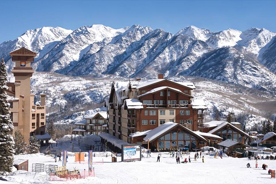 A snow-covered mountain resort with a ski lift, buildings, and a clear blue sky.