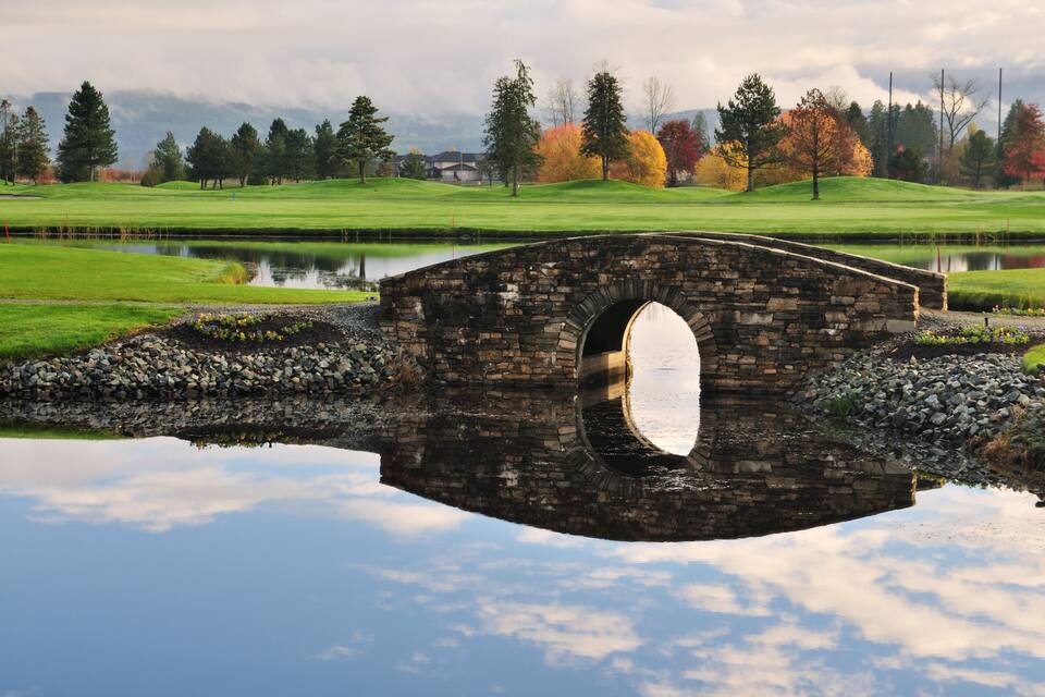Un pont de pedra sobre un estany tranquil amb un camp de golf i arbres d'hivern a la part posterior.