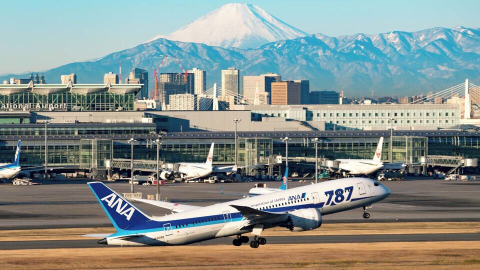 An ANA airplane on a runway with a cityscape and a mountain in the background.