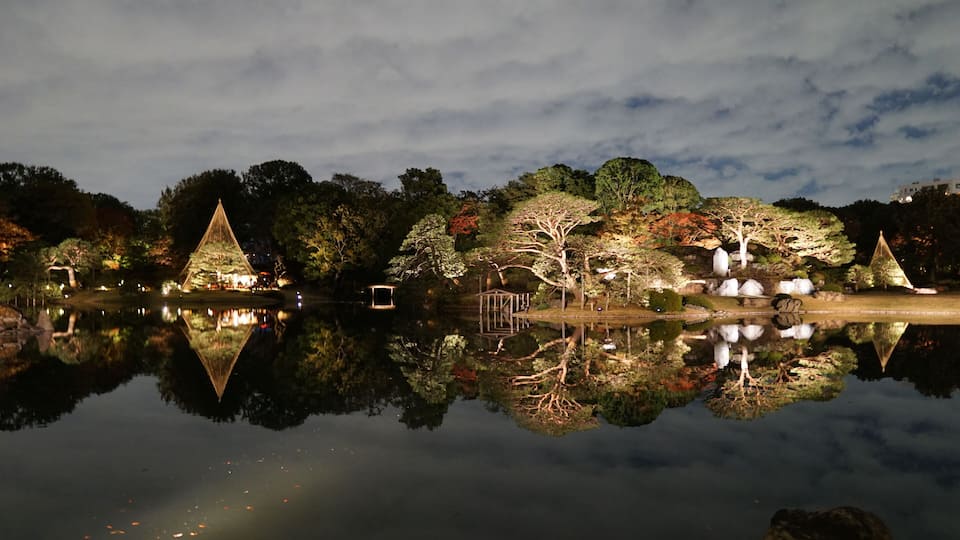 Une scène nocturne paisible, avec un étang aux reflets argentés, des arbres illuminés et des structures géométriques.
