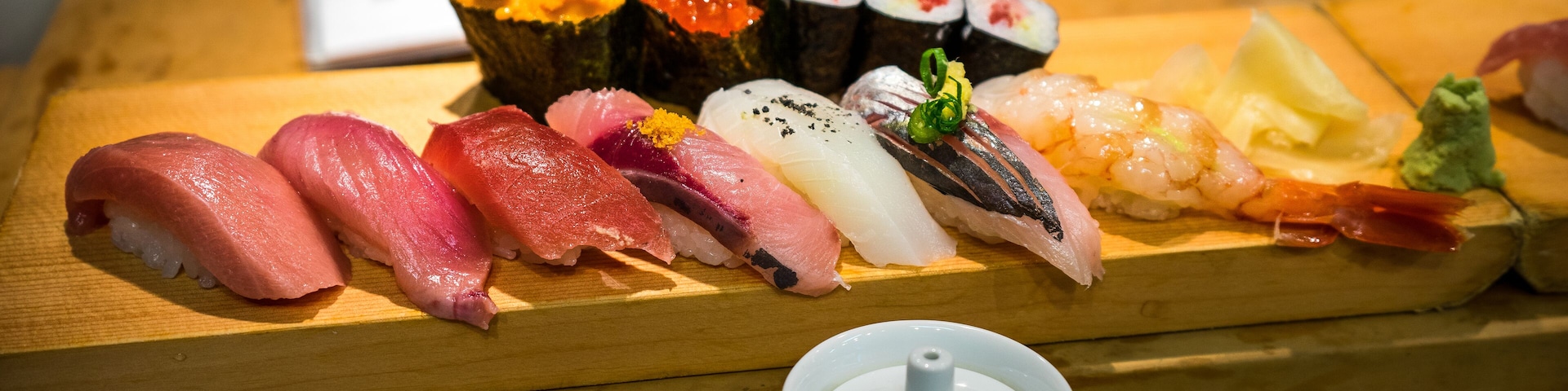 Une table en bois sur laquelle sont disposés différents types de sushis et de sashimis, notamment du saumon, du thon et des crevettes, ainsi qu’une théière blanche.