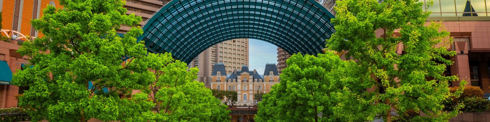 A pedestrian pathway lined with trees, a large arch structure, and modern buildings in the background.