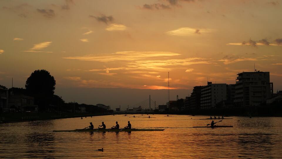 A sunset over a river with people rowing canoes, buildings on the riverbank, and a bridge in the distance.