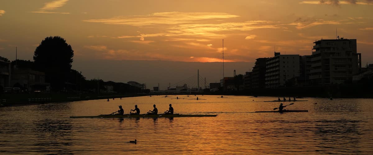A sunset over a river with people rowing canoes, buildings on the riverbank, and a bridge in the distance.