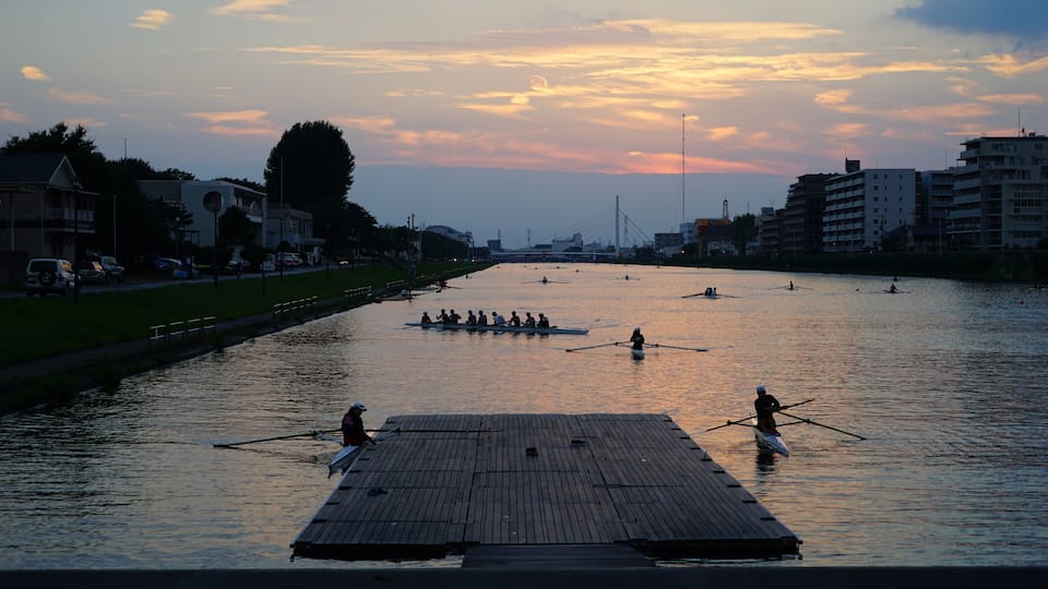 A sunset over a river with rowing boats, a wooden dock, and buildings along the riverbank.