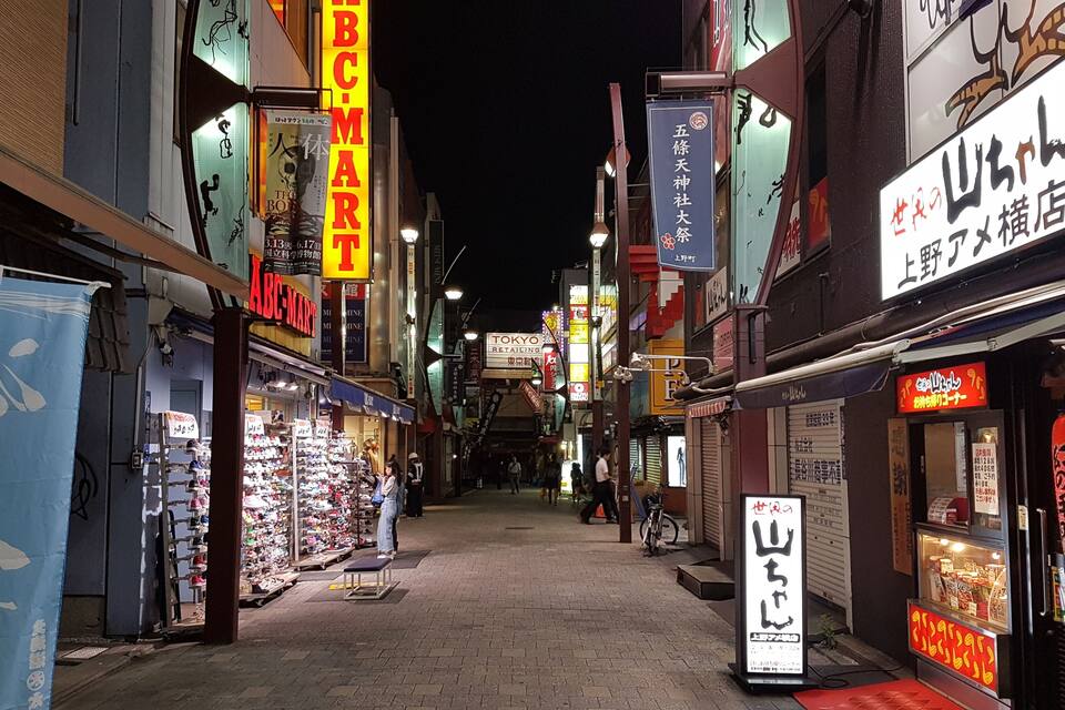 Uma rua estreita repleta de lojas e placas, incluindo uma loja de sapatos e um restaurante.