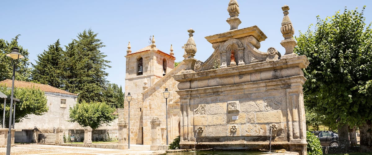 A historic fountain with ornate stone carvings, a church with a bell tower, and a cobblestone plaza.