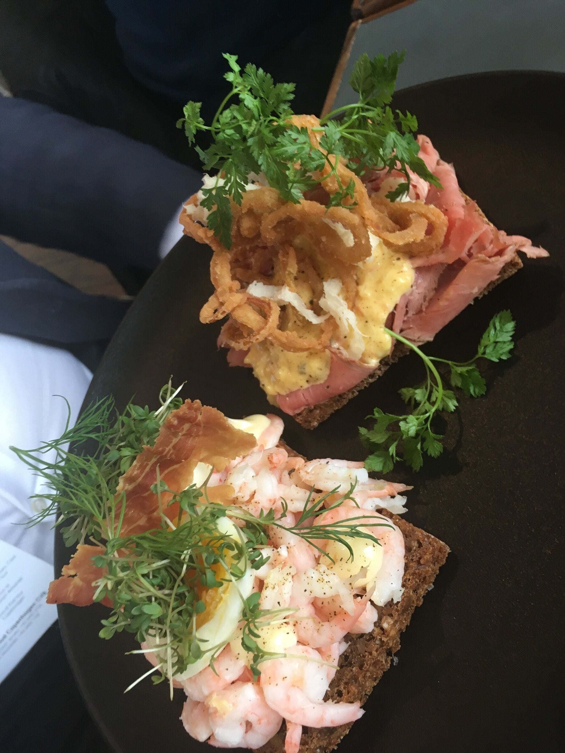 Two open-faced sandwiches with shrimp, eggs, and greens on dark bread.