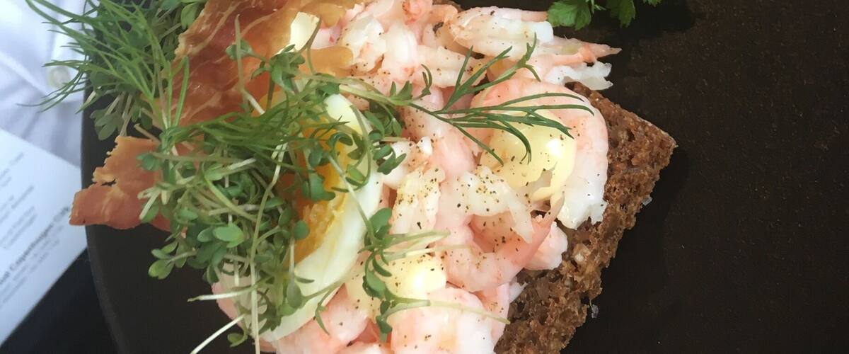 Two open-faced sandwiches with shrimp, eggs, and greens on dark bread.