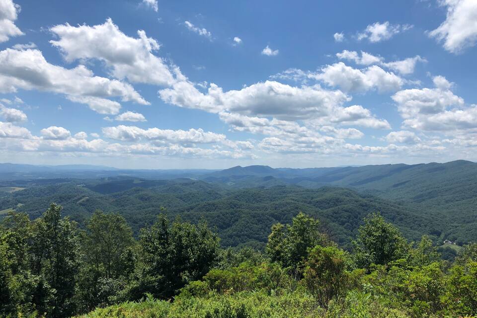 A mountainous landscape with dense green forests and a clear blue sky with scattered clouds.