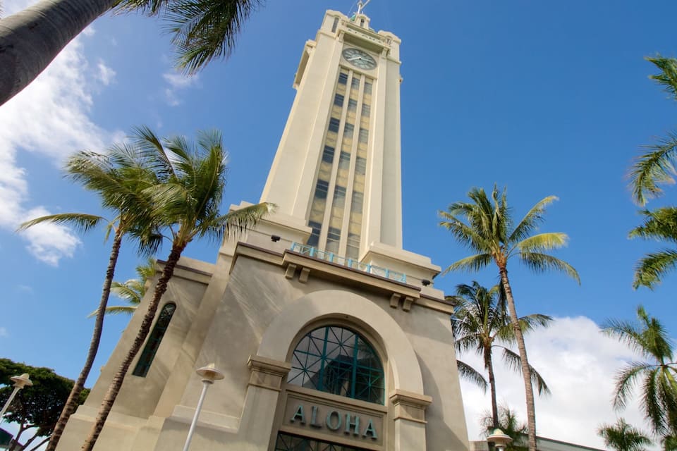 A tall clock tower with the word "ALOHA" on its base, surrounded by palm trees.