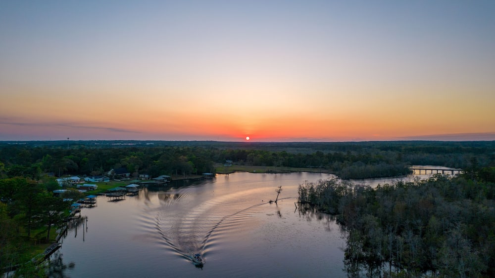 Ein Sonnenuntergang über einem ruhigen See, mit einem Boot, das Wellen verursacht, umgeben von Bäumen und einer entfernten Brücke.
