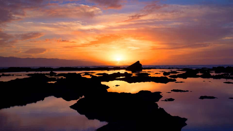 A sunset over a rocky shoreline with calm water reflecting the sky.