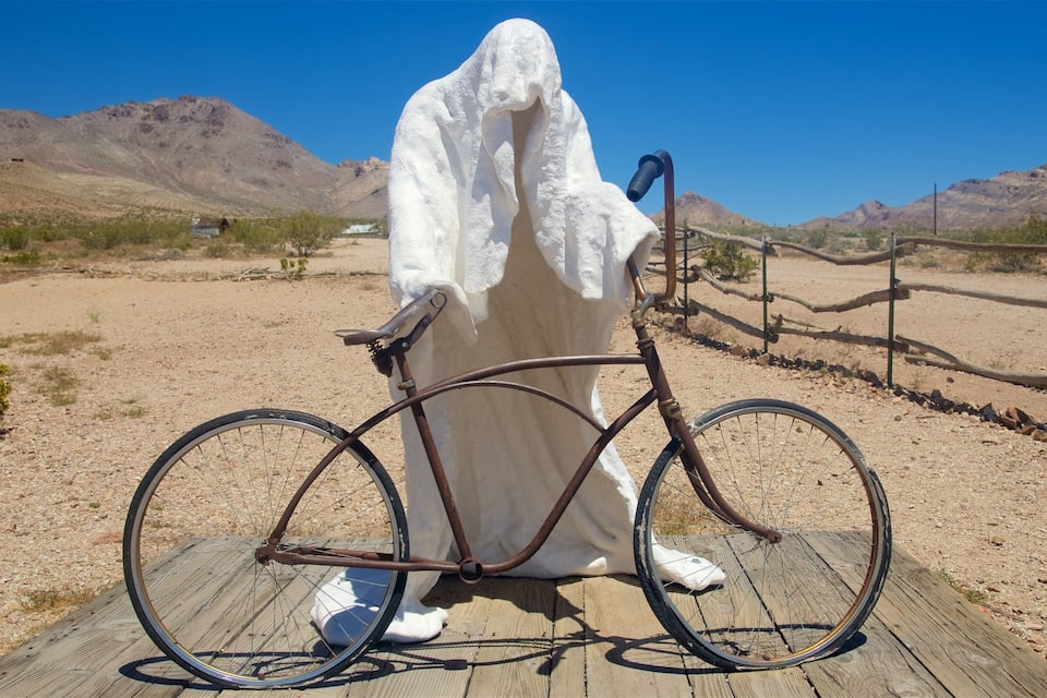Uma figura fantasmagórica branca sentada em uma plataforma de madeira com uma bicicleta em uma paisagem desértica.