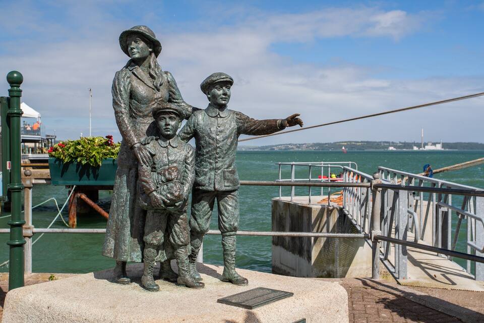 A bronze statue of a woman and child on a pier with a ship in the background.