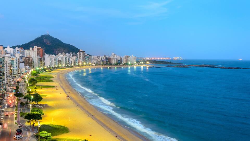 A coastal cityscape with a prominent mountain, a wide beach, and a dense urban area with high-rise buildings.