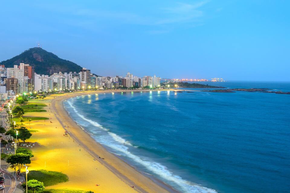 A coastal cityscape with a prominent mountain, a wide beach, and a dense urban area with high-rise buildings.