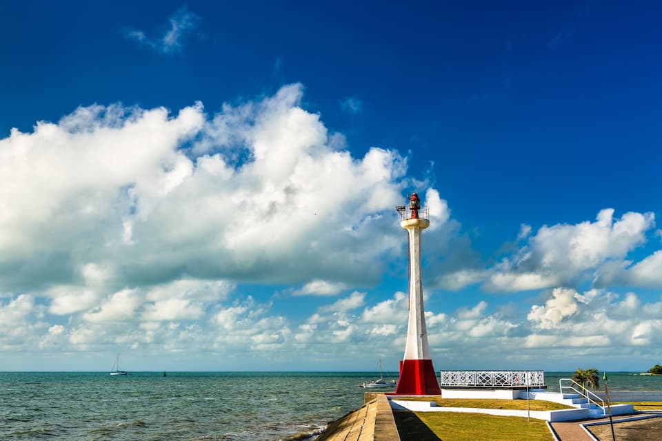 Um farol com base vermelha e branca, uma passarela e vista do oceano com um veleiro ao longe.