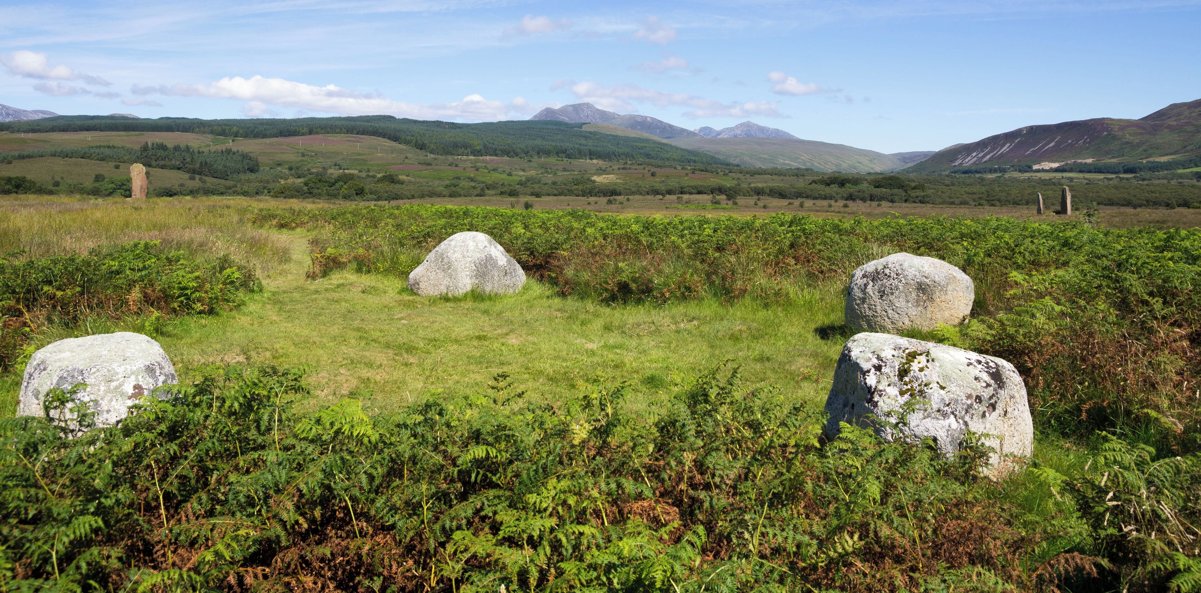 Circle 4 at Machrie Moor. Four rounded granite blocks, just less than 1m high. In the middle distance on the left is the large stone of circle 3 and on the right are the three large stones of circle 2. In the far distance are the mountains of the north of Arran, consisting of Beinn Tarsuinn and Goat Fell.