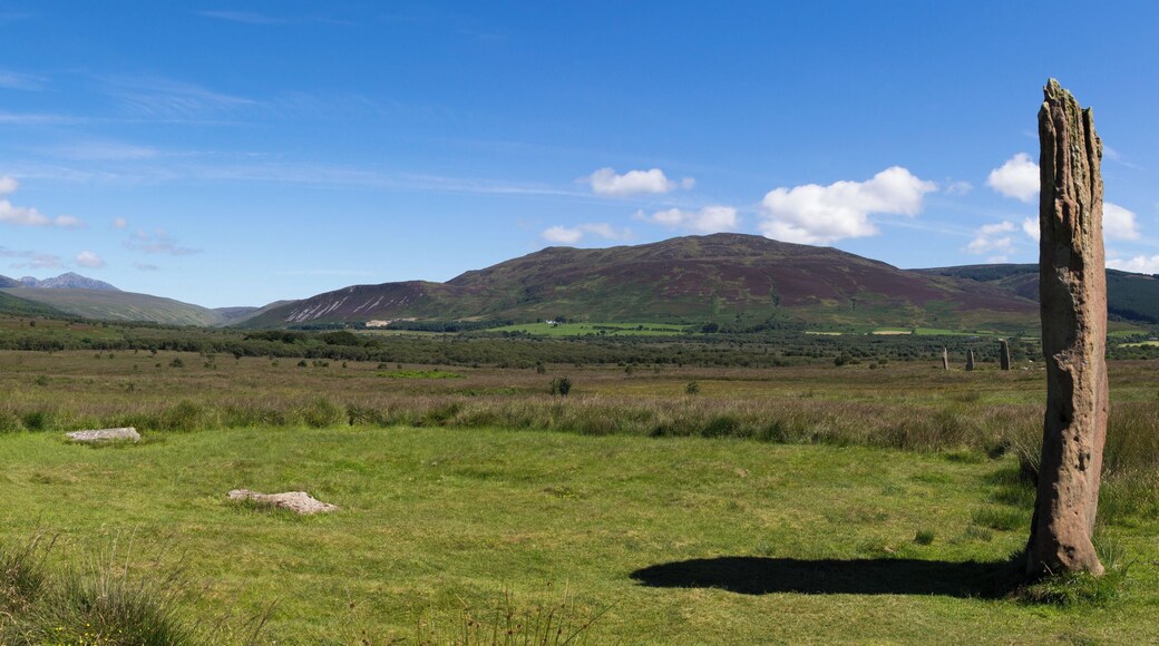 Circle 3 at Machrie Moor. One remaining stone standing 4.3m tall. The stumps of three others remain. In the middle distance to the right are the three large stones of circle 2. In the far distance to the left are the mountains of the north of Arran, consisting of Beinn Tarsuinn and Goat Fell.