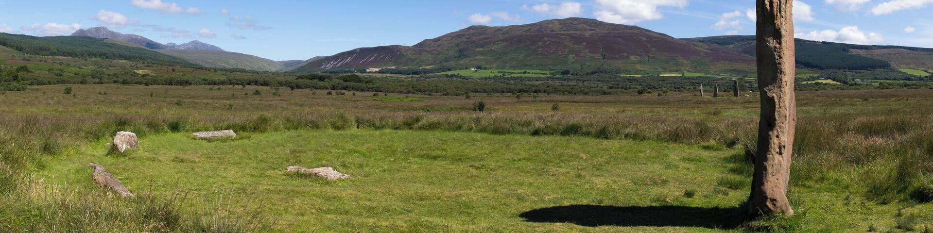 Circle 3 at Machrie Moor. One remaining stone standing 4.3m tall. The stumps of three others remain. In the middle distance to the right are the three large stones of circle 2. In the far distance to the left are the mountains of the north of Arran, consisting of Beinn Tarsuinn and Goat Fell.