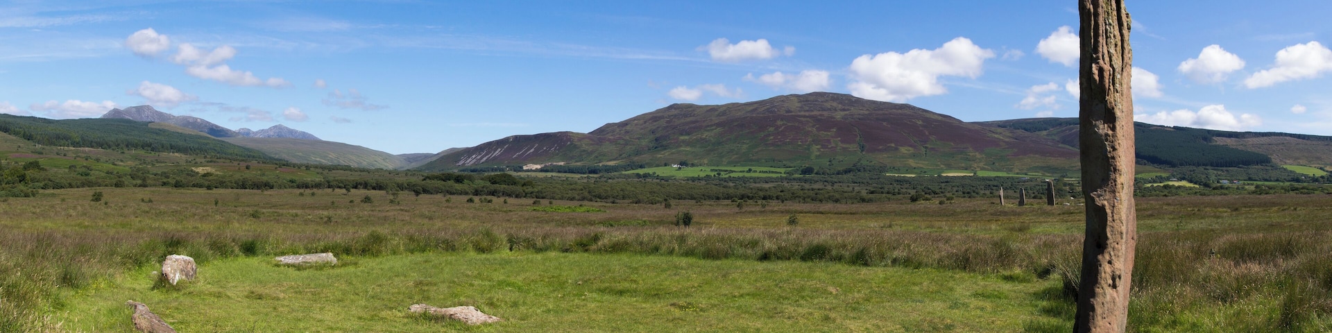 Circle 3 at Machrie Moor. One remaining stone standing 4.3m tall. The stumps of three others remain. In the middle distance to the right are the three large stones of circle 2. In the far distance to the left are the mountains of the north of Arran, consisting of Beinn Tarsuinn and Goat Fell.