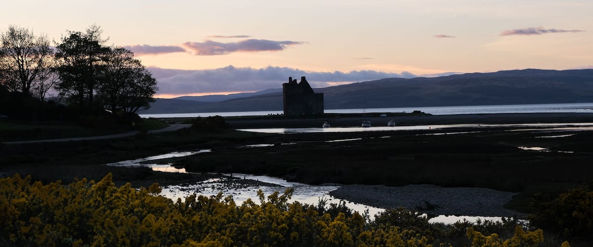 A coastal landscape with a castle, a river, and a mountainous backdrop.