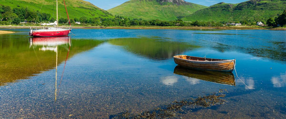A calm lake with a red sailboat and a wooden rowboat, surrounded by green hills and a clear blue sky.