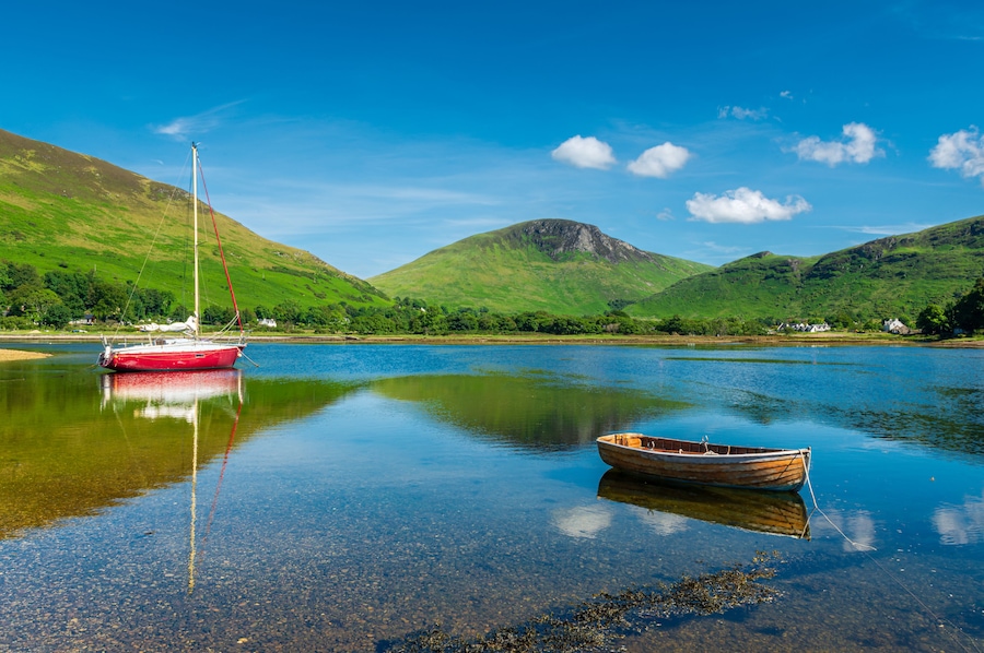 A calm lake with a red sailboat and a wooden rowboat, surrounded by green hills and a clear blue sky.