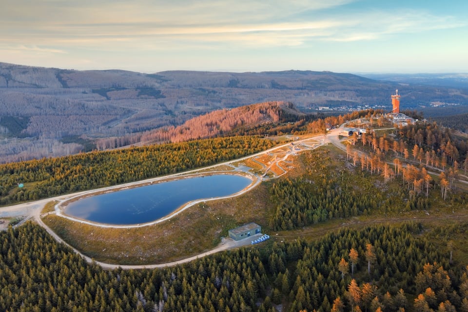 An aerial view of a lake surrounded by dense forests, a small building, and a tall tower.