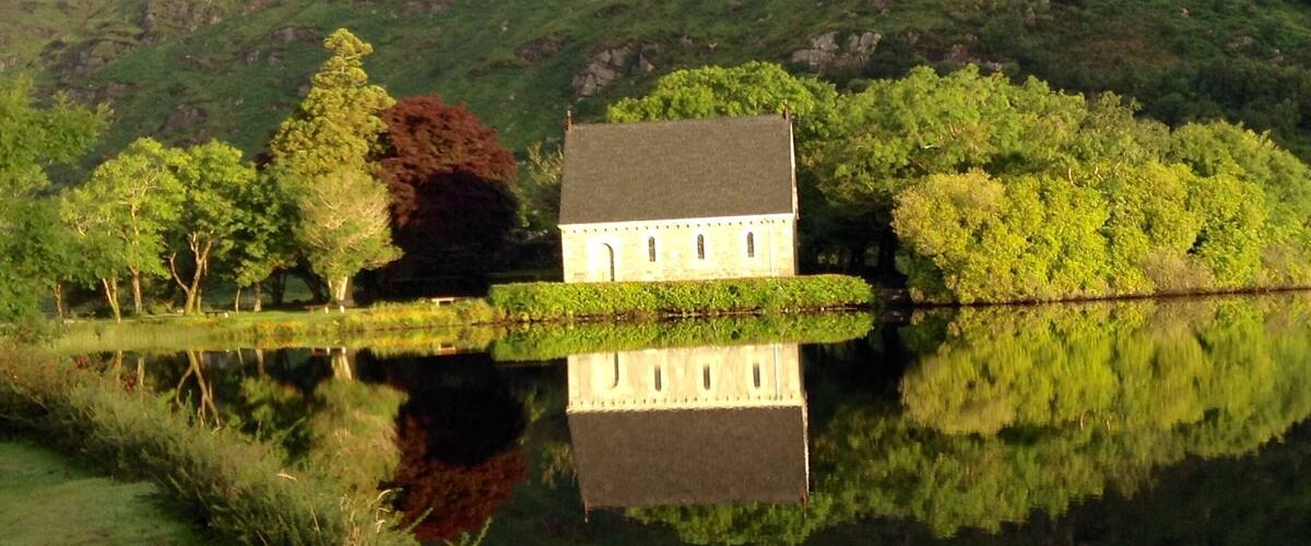 A house by a calm lake with a reflection, surrounded by lush greenery and hills.